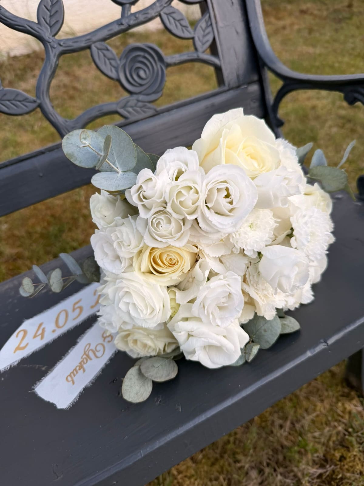 Brautstrauß aus weißen Rosen, Lisianthus und Eukalyptus, abgelegt auf einer dunkelgrauen Gartenbank, daneben ein Satinband mit aufgedrucktem Hochzeitsdatum